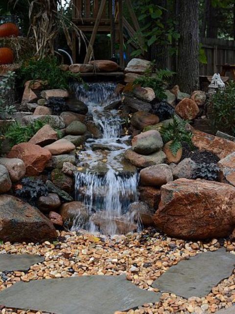 a cool backyard waterfall styled in a natural way, with large rocks and greenery around, with pebbles on the ground is lovely for outdoors