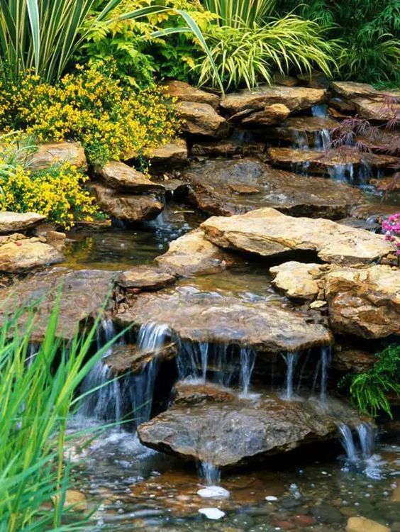 a cool backyard waterfall with rocks placed on a slope, with greenery and blooms is a lovely idea that feels Alpine