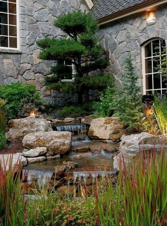 a front yard water feature with large rocks and herbs all around and a large tree over it