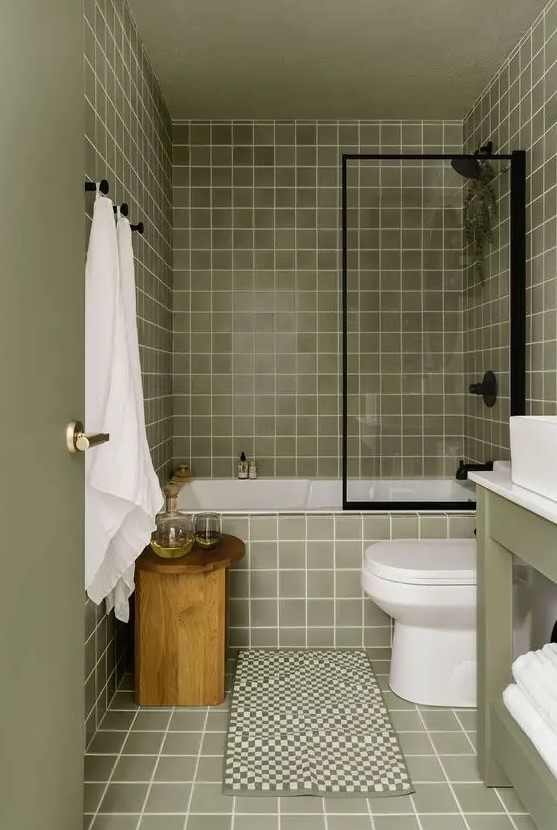 a light green bathroom clad with square tiles, a green vanity, a wooden stool, black fixtures and white appliances