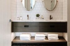 36 a mid-century modern bathroom clad with white square tiles, a black vanity and a shelf, oval mirrors, towels and a printed rug