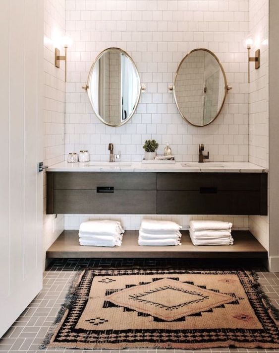 a mid-century modern bathroom clad with white square tiles, a black vanity and a shelf, oval mirrors, towels and a printed rug