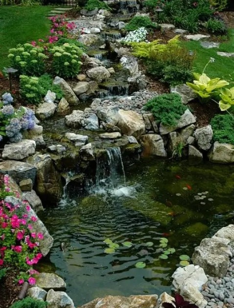 a lovely garden waterfall placed on a slope, composed of rocks and surrounded with blooms and greenery is a cool idea to add an Alpine touch to the space