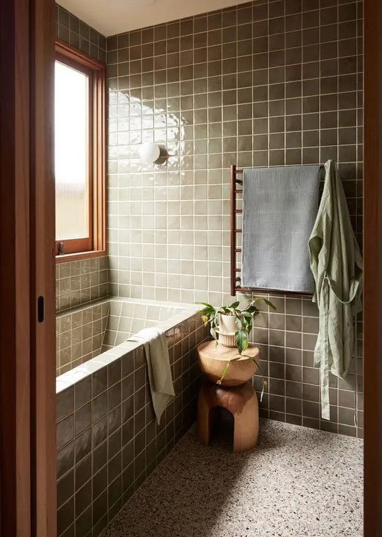 a modern bathroom with a terrazzo floor, green square tiles and a tub clad with tiles, timber and stained wood plus a wooden stool