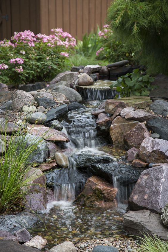 a pretty all-natural waterfall composed of large and small rocks, with greenery and pink blooms is a cool idea for a garden