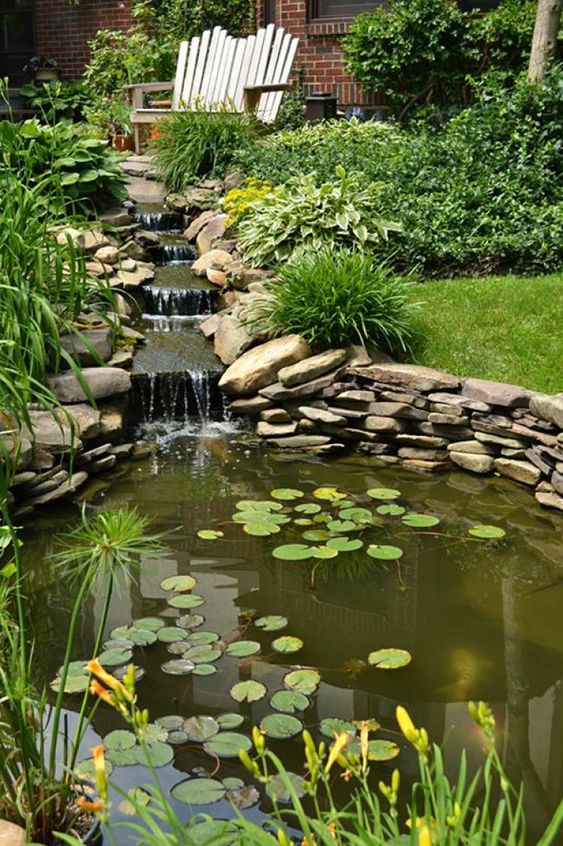 a relaxed garden with a green lawn, some bushes and a descending waterfall clad with rocks, a wooden bench and some decor