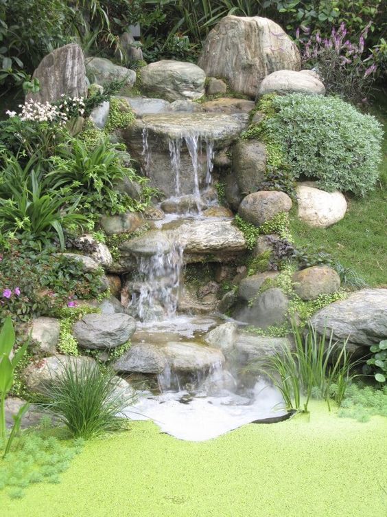 a rustic backyard waterfall with large rocks, greenery and blooms and a pond with green grass is a cool idea for a rustic garden