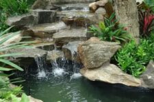 49 a small all-natural waterfall made of oversized rocks, with a tree, some greenery and blooms around and a deck to sit and watch it