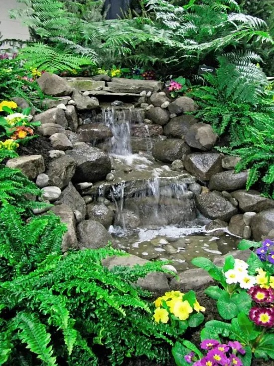 an Alpine-style waterfall made of rocks, with greenery and bright blooms around is a cool and bright way to add interest to your garden