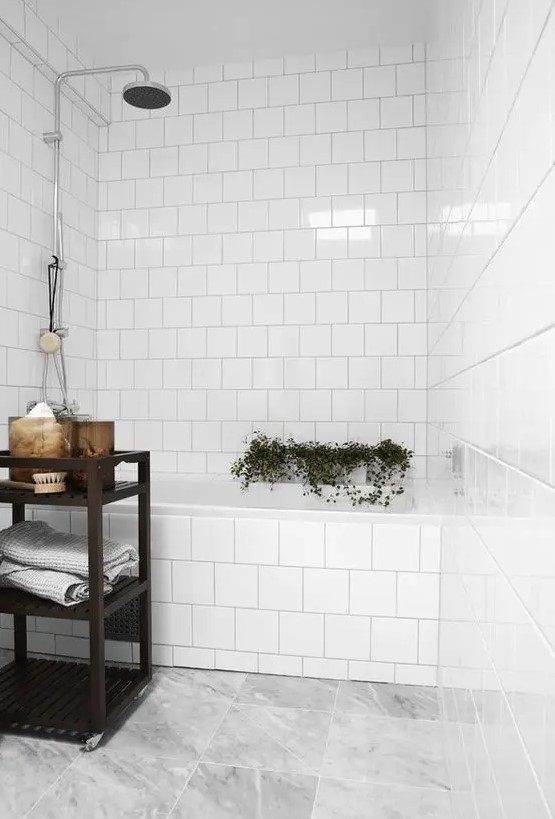 a simple white bathroom with white square tiles and a grey marble tile floor, a black shelving unit, some greenery and decor