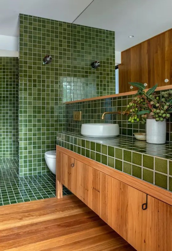 a welcoming bathroom with green square tiles and a stained floor and vanity, white applinaces and brass fixtures and a large mirror