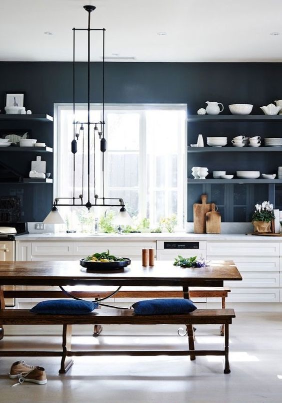a bold kitchen with navy walls and matching open shelves, white lower cabinets, a stained dining set with benches and a unique lamp