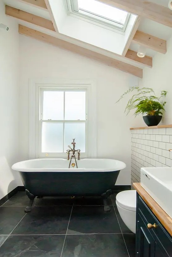 a chic attic bathroom with a window and a skylight, black large scale tiles and white subway ones, a black vanity and a black tub