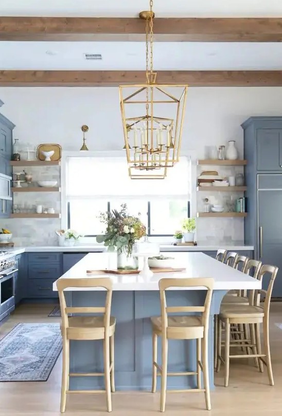 a chic coastal kitchen with ocean blue shaker cabinets, marble tiles, a large kitchen island with white countertops, wooden stools and gold pendant lamps