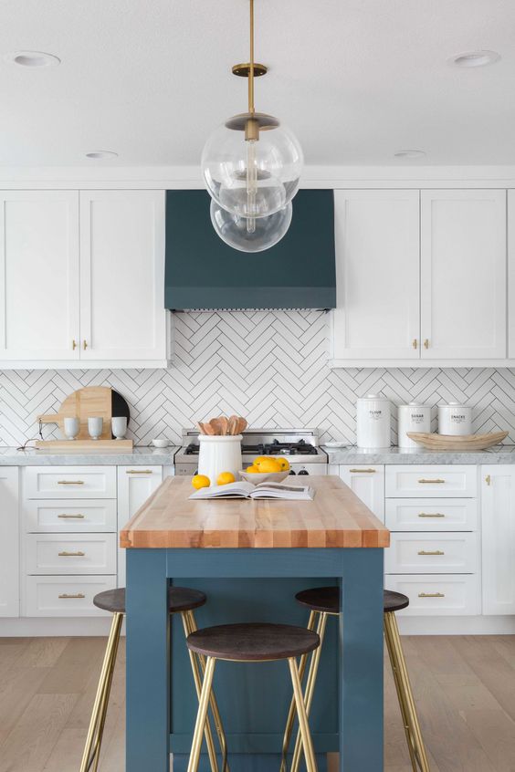 a chic farmhouse kitchen with white shaker cabinets, a herringbone tile backsplash, a blue kitchen island and a hood
