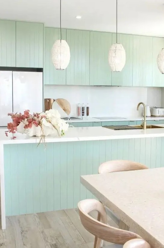 a chic mint green slatted kitchen with a white backsplash and countertops, cool pendant lamps, a terrazzo table and stained chairs