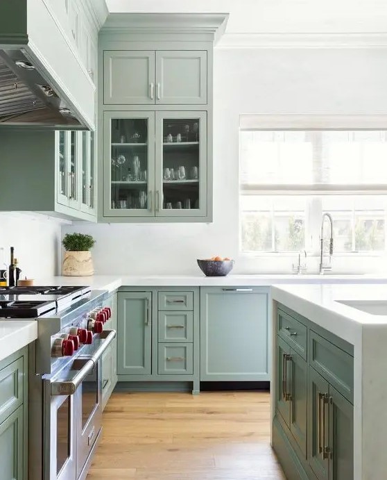 a classic sage green shaker style kitchen with glass front cabinets, white stone countertops and backsplash and brass handles