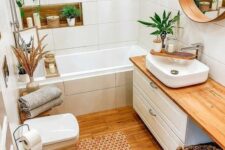 a cool small bathroom clad with large scale tiles, a white vanity, a round mirror, potted greenery and candles and a basket