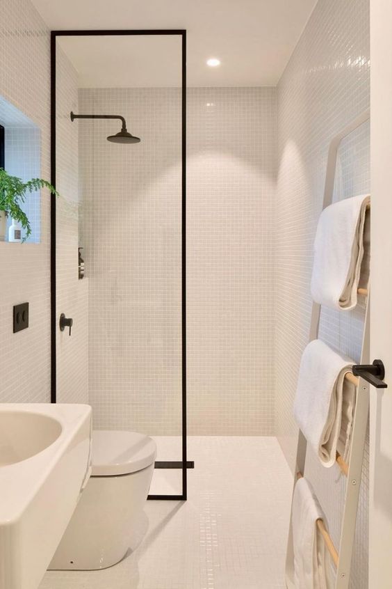 a cozy neutral bathroom with small tiles, a shower space with black framing, a sink and a toilet, a ladder with towels