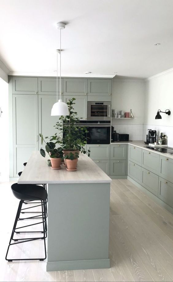a lovely and soothing sage green kitchen with shaker cabinets, a kitchen island, black stools, pendant lamps and a white tile backsplash