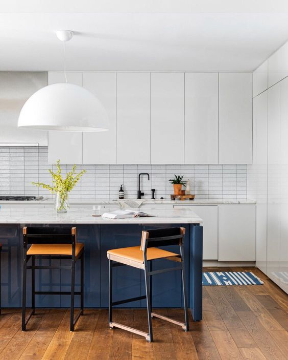 a minimal kitchen with sleek white cabinets and a skinny tile backsplash, a navy kitchen island and amber stools