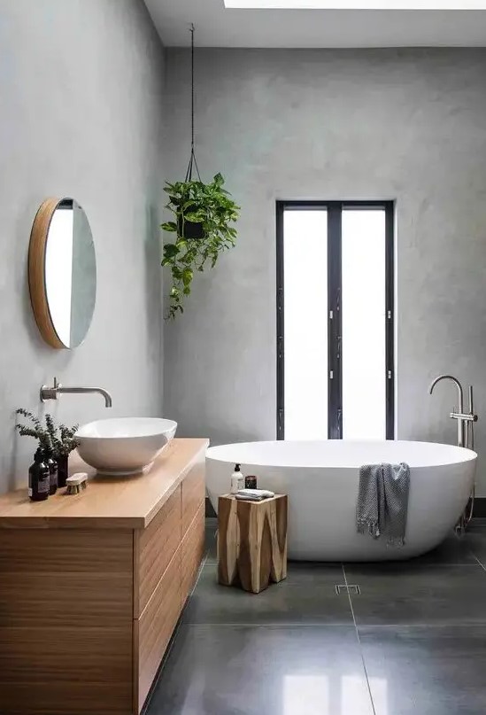 a minimalist grey bathroom with concrete walls and large scale tiles on the floor, a stained vanity, an oval tub, potted greenery and a window