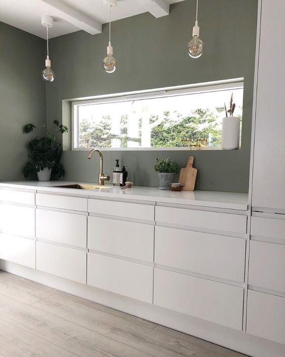 a minimalist kitchen with green walls and sleek white cabinets, pendant bulbs and a window instead of a backsplash