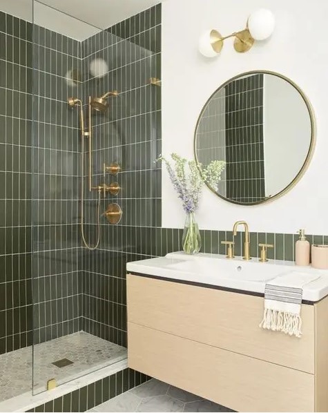 a modern bathroom with green skinny tiles, a floating vanity, a round mirror, gold and brass fixtures is cool