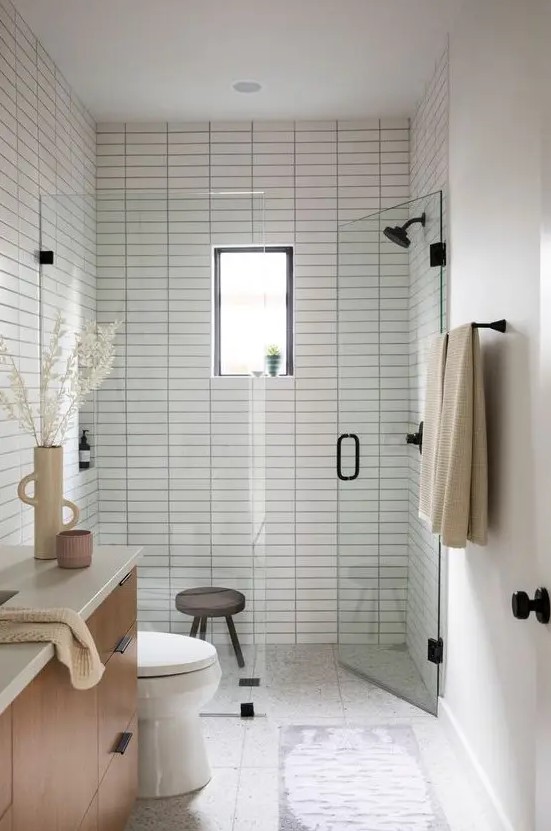 a peaceful and welcoming small bathroom clad with white skinny tiles, with a chic vanity, black fixtures and a small window in the shower