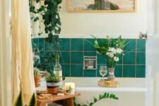 a pretty eclectic bathroom with green tiles, a neutral tub, a wooden stool, potted plants, a lovely artwork and neutral textiles