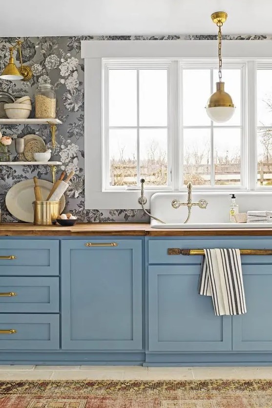 a refined kitchen with grey floral print wallpaper, blue shaker cabinets, butcherblock countertops, gold handles and a gold pendant lamp