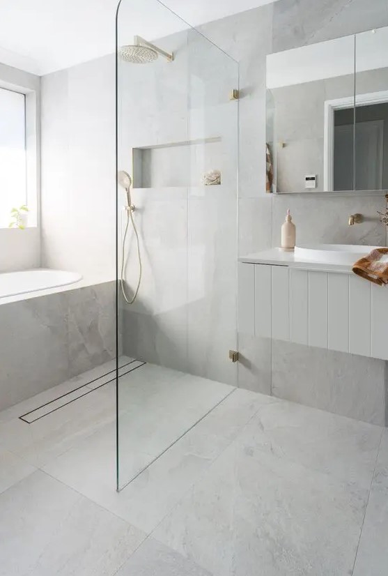 a serene minimalist bathroom with grey large scale tiles, a tub clad with tiles, a shower space, a vanity and brass touches