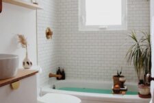 a small and cool boho bathroom with white subway tiles, a tub, a white vanity, open shelves, potted greenery, a rug and candles