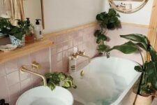 a small boho bathroom with pink tiles, an oval tub, a vanity with a sink, some potted plants and a shelf
