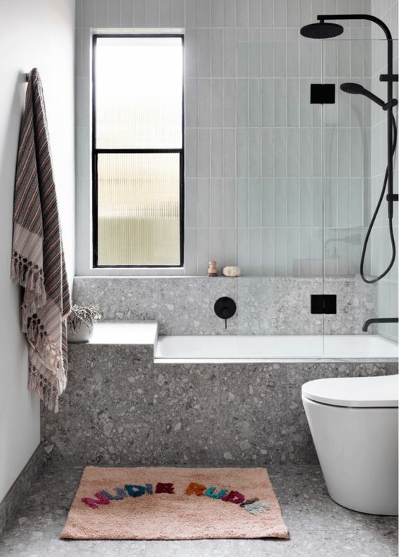 a small contemporary bathroom clad with pale blue tiles and grey terrazzo, a tub, a toilet, black fixtures and a window with reeded glass