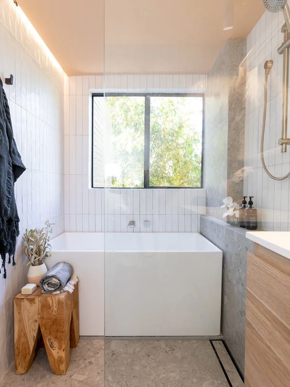 a small contemporary bathroom with a window, a square tub, white tiles and terrazzo, a vanity, a wooden stool and lights
