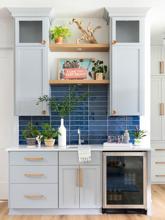 a small light grey kitchen with shaker cabinets and a navy skinny tile backsplash, open shelves and a drink cooler plus potted plants