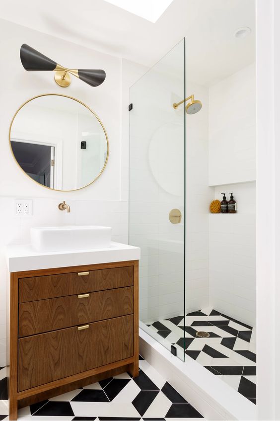 a small modern bathroom with a shower space, a tiled black and white floor, a stained vanity, a black sconce and a round mirror