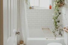 a small neutral boho bathroom clad with white subway and penny tiles, a bathtub, a toilet, some potted greenery and a curtain