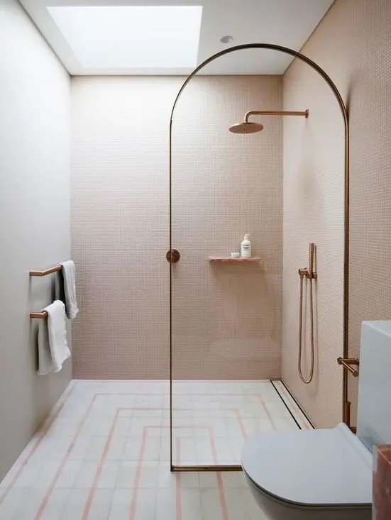 a small unique bathroom clad with blush mini tiles, with neutral and pink ones on the floor and a skylight over the shower plus brass touches