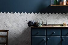 a stylish kitchen wiht navy walls and matching cabinets, a grey tile floor and grey marble herringbone tiles, an open shelf and a pendant bulb