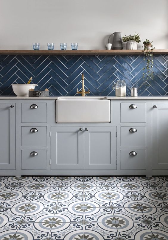 a stylish kitchen with dove grey shaker style cabinets, printed tiles on the floor, a navy herringbone tile backsplash and an open shelf