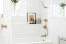a stylish neutral bathroom with a tub and a shelf over it, a white vanity, a toilet and brass fixtures