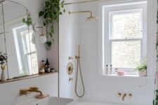 a tiny and cool bathroom wiht a bathtub next to the window, a wall-mounted sink, a pink pouf, gold fixtures and some greenery