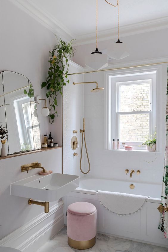 a tiny and cool bathroom wiht a bathtub next to the window, a wall-mounted sink, a pink pouf, gold fixtures and some greenery