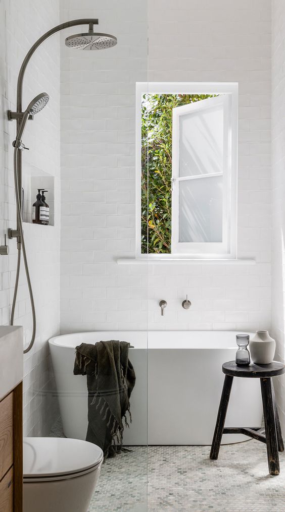 a tiny contemporary bathroom with a tub next to the window, a vanity, a toilet and a black stool as a stand