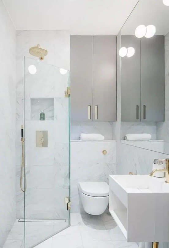 a tiny minimalist bathroom with white marble tiles, sleek grey cabinets, gilded touches and a triangle shower space