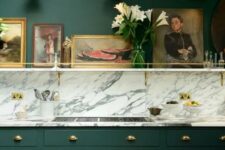 a vintage hunter green kitchen with a white marble backsplash and countertops, a shelf with artworks is chic