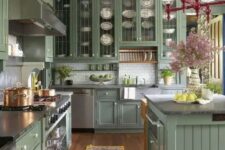 a vintage sage green kitchen with glass and usual cabinets, grey stone counterops, a white tile backsplash and a vintage chandelier
