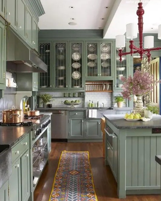 a vintage sage green kitchen with glass and usual cabinets, grey stone counterops, a white tile backsplash and a vintage chandelier